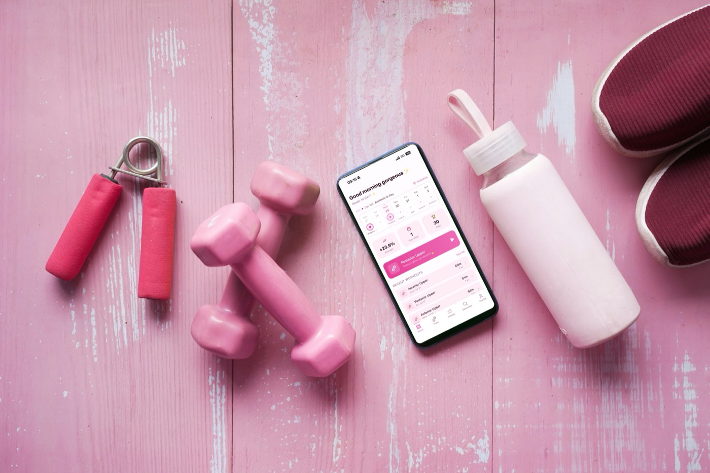 Pink flat-lay of workout gear — dumbbells, a hand gripper, a water bottle, and a phone showing the NorseLift workout tracker app home screen